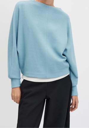 Frau trägt einen hellblauen Pullover mit langen Ärmeln über einem weißen Unterhemd und schwarze Hosen, steht vor einem einfarbigen Hintergrund.