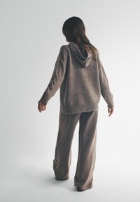 Kobieta zwrócona tyłem, ubrana w luźną brązową dzianinową bluzę z kapturem i pasujące szerokie spodnie, stojąca na jednolitym jasnym tle.