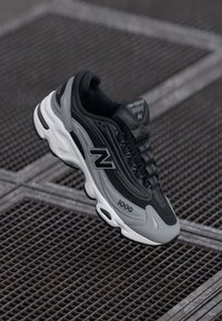 Black and gray athletic shoe with mesh upper, textured design, prominent "N" logo, padded collar, and white midsole detailing.