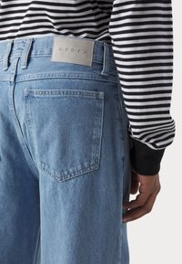 Ljusblå denim jeans med rak passform, som har en bakficka och en grå läderlapp på midjebandet. Randig svart och vit topp synlig.