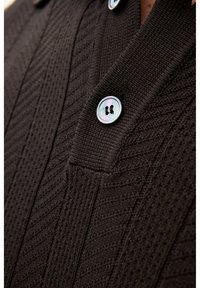 Nahaufnahme eines dunkelbraunen, gestrickten Cardigans mit strukturierten Chevron-Mustern und einem glänzenden Vierlochknopf, der vorne befestigt ist.