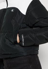 Černobílá zimní bunda s logem Calvin Klein Jeans na rukávu a emblémem cK na hrudi, ruka v postranní kapse.