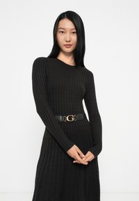 Abito nero a costine con maniche lunghe, caratterizzato da una silhouette aderente e una cintura nera con dettagli dorati in vita. Texture liscia.