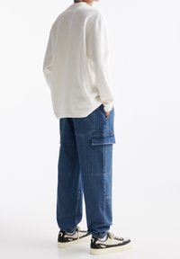 Person som bär en vit långärmad skjorta, blå denim cargo-byxor med sidofickor och vita sneakers med svarta detaljer som står mot en vit bakgrund.