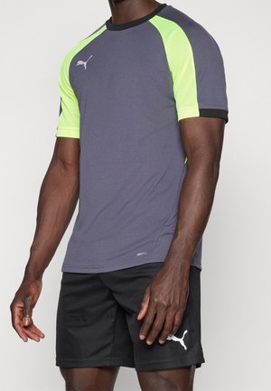 Homme portant un t-shirt de sport Puma gris et jaune fluo avec un short Puma noir, debout devant un fond uni.