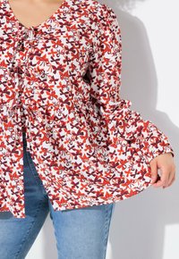 Bluse med rød og lilla blomstermønster, V-hals, knyttet foran og lange puffede ærmer, lavet af let stof, båret over blå jeans.