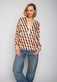 Bluzka z długim rękawem w lekkim, błyszczącym materiale z pomarańczowym, granatowym i kremowym geometrycznym wzorem. Dekolt Henley z delikatnym wykończeniem.