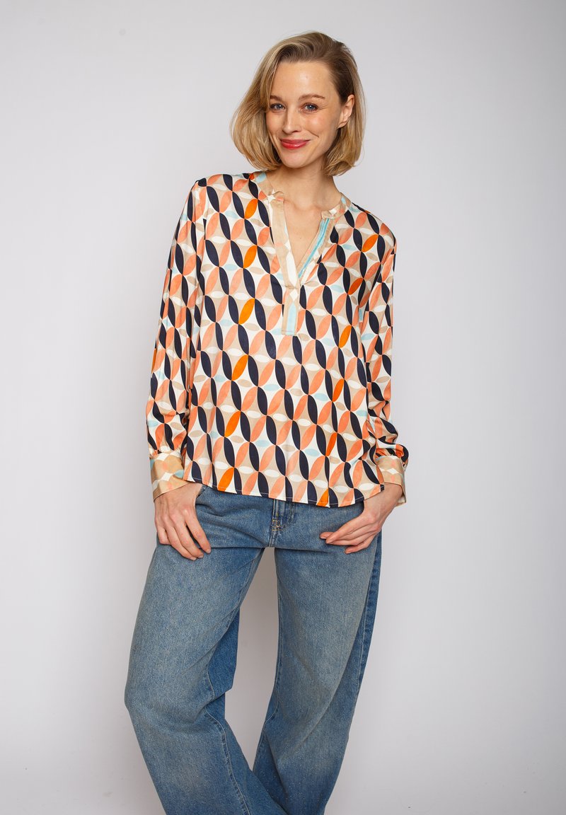 Bluzka z długim rękawem w lekkim, błyszczącym materiale z pomarańczowym, granatowym i kremowym geometrycznym wzorem. Dekolt Henley z delikatnym wykończeniem.