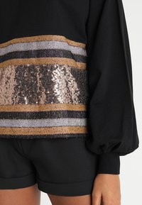 Maglietta nera a maniche lunghe con righe orizzontali in paillettes dorate, argentate e nere. Materiale morbido con maniche a sbuffo.
