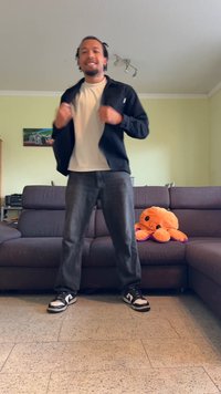 Une personne portant une veste noire sur une chemise blanche, un jean gris et des baskets noires. Un jouet en peluche d'octopus orange est posé sur un canapé marron à l'arrière-plan.