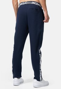 Marineblauwe sweatpants met een logo-broekband, een zijzak en witte merkletters langs het been, gemaakt van een zachte, gestructureerde stof.