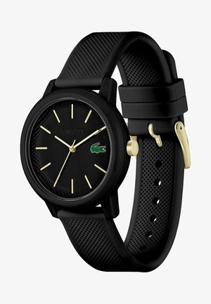 Orologio in gomma nera con cinturino testurizzato, indicatori delle ore dorati e un quadrante nero con il logo verde di Lacoste. Design del case circolare.
