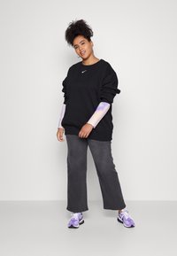 Črna sweatshirt s Nike logotipom, rokavi s vijoličnim in belim vzorcem, v kombinaciji s sivimi širokimi hlačami in vijoličnimi športnimi copati.