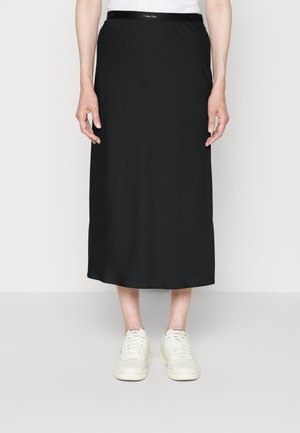Jupe midi noire Calvin Klein avec une texture lisse, une taille élastique et une coupe droite. Portée avec des sneakers blanches Reebok.