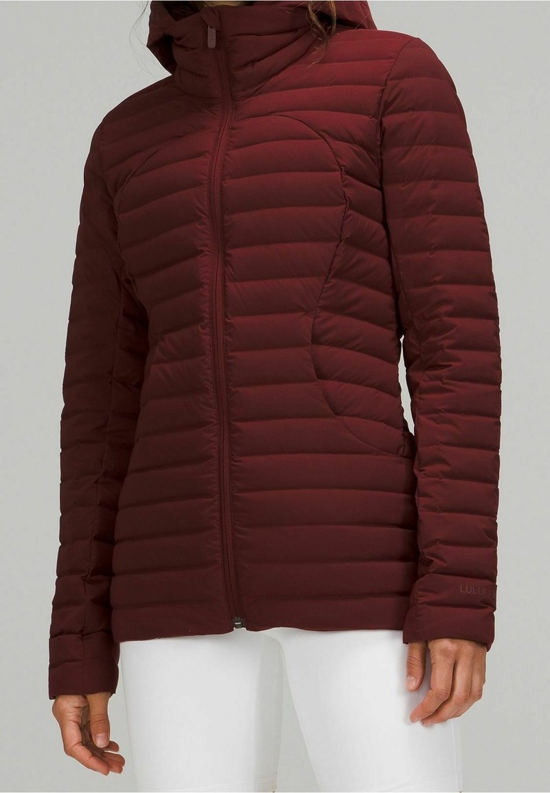 Kobieta ubrana w bordową pikowaną kurtkę z zamkiem z przodu oraz białe szorty na jednolitym tle.