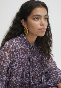 Lila blus med blommönster, långa ärmar, veckdetaljer och rund krage. Åtföljd av stora, texturerade guldörhängen.