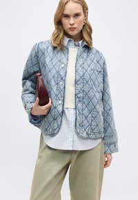 Γυναίκα με ξανθά μαλλιά που φοράει μπλε καπιτονέ τζιν μπουφάν πάνω από ριγέ πουκάμισο και μπεζ παντελόνια, κρατώντας σκούρο κόκκινο τσάντα φάκελο με υφή.