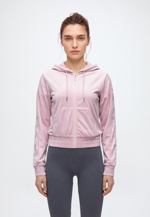 Une femme porte un sweat à capuche rose pâle zippé, avec des bordures à motifs sur les manches, et un legging gris foncé sur un fond uni.