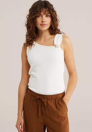 Žena s kučeravými hnedými vlasmi, ktorá nosí biely tielkový top s brošňou v tvare morskej hviezdice a hnedé nohavice so šnúrkou, stojaca pred béžovým pozadím.