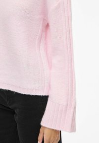 Jasnoróżowy sweter z dzianiny z prążkowymi mankietami i bocznymi detalami, o luźnym kroju i miękkiej teksturze. W zestawie z czarnymi spodniami.