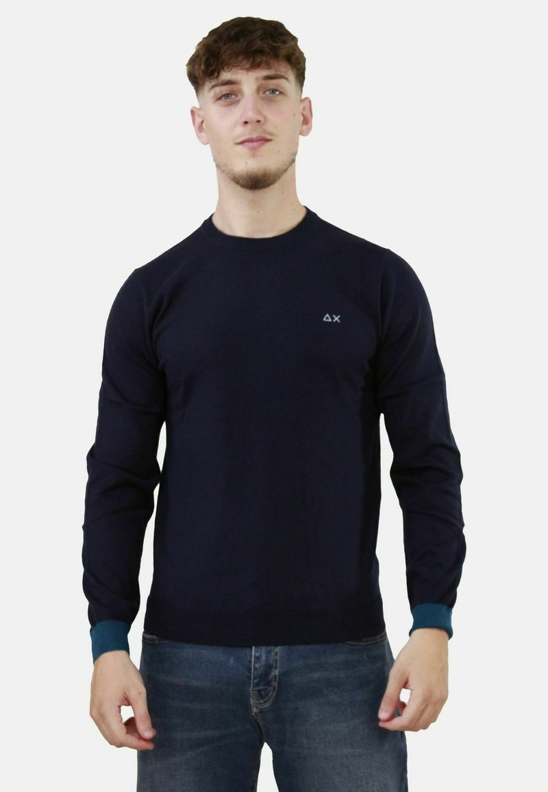 Maglione lungo a maniche con polsini a costine di colore blu navy, con scollatura rotonda e un piccolo logo sul petto. Indossato sopra a jeans chiari.
