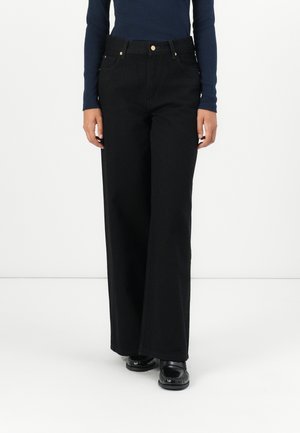 Persoon draagt zwarte broek met hoge taille en wijde pijpen, met steekzakken aan de voorkant en zwarte schoenen, staand tegen een effen witte achtergrond.