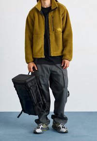 Giacca in pile di colore verde oliva con tasca sul petto con zip e cuciture piatte; abbinata a pantaloni cargo scuri e scarpe da ginnastica grigie. Porta uno zaino nero.