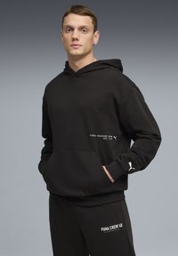 Sweat à capuche noir avec une poche kangourou, arborant des logos et du texte blancs. Tissu doux, coupe décontractée, capuche réglable et poignets côtelés.