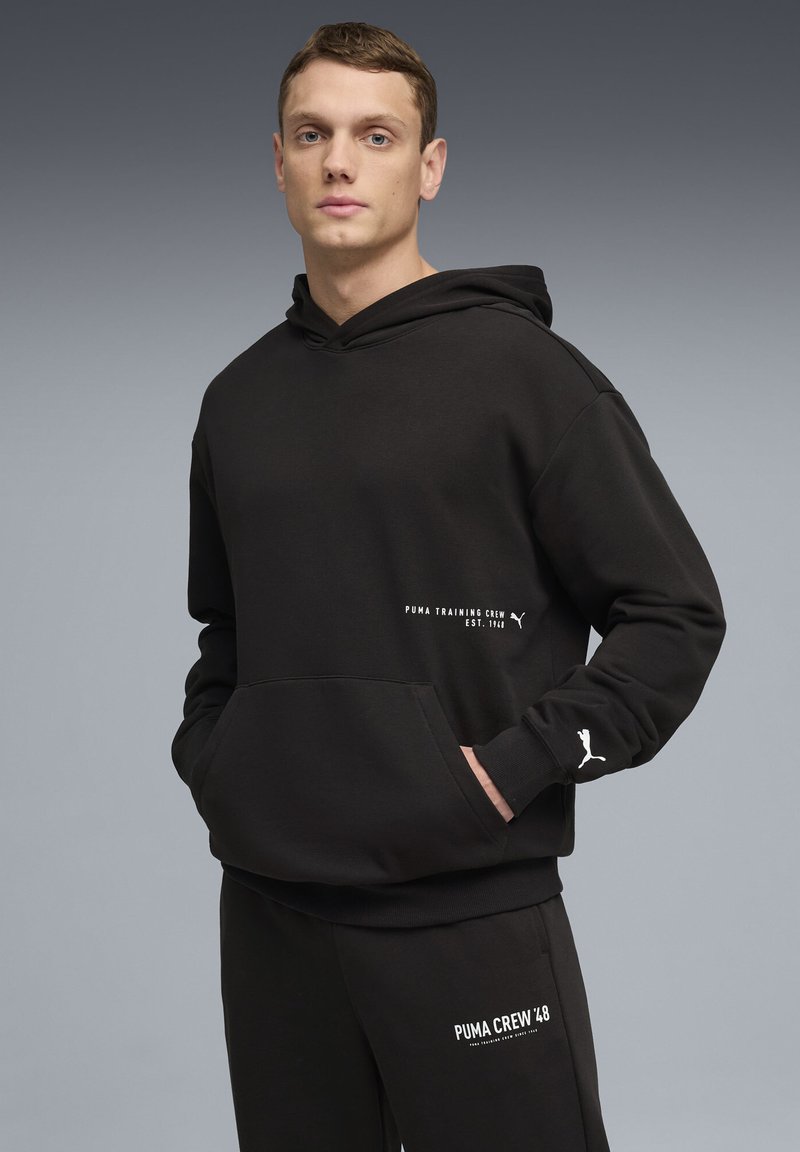 Sweat à capuche noir avec une poche kangourou, arborant des logos et du texte blancs. Tissu doux, coupe décontractée, capuche réglable et poignets côtelés.