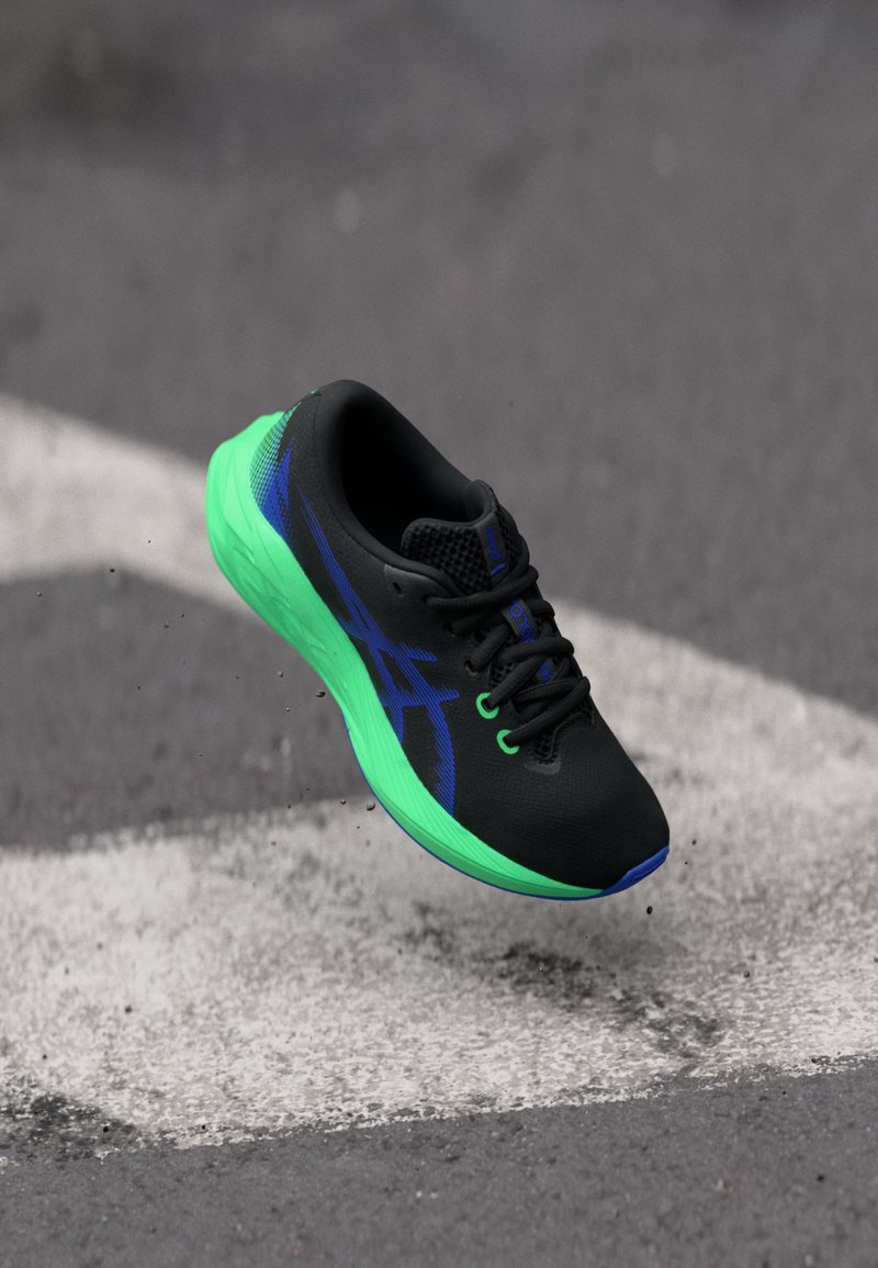 Chaussure de sport noire avec semelle verte, accents bleus, tige en textile léger et système de laçage, planant au-dessus d'une surface grise.