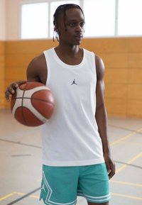 Bílý sportovní tílko s síťovou strukturou, s malým černým logem. Spárováno s aqua kraťasy s bílými akcenty. Drží basketbal.