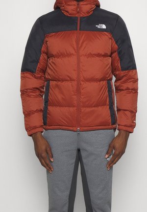 Homme portant une doudoune rouge rouille et noire avec capuche, fermeture éclair avant, et un pantalon gris matelassé avec une bande noire et un logo sur la cuisse.