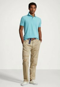 Young man wearing a teal polo shirt, beige ripped pants with a striped belt, and white sneakers, standing with hands in pockets.