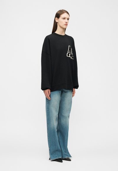 Fiatal személy áll, fekete, túlméretezett pulóvert visel fehér logóval és világoskék, széles szárú farmernadrágot, fehér háttér előtt.