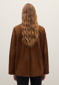 Brown suede jacket with long sleeves and a relaxed fit; visible stitching along the shoulders and a smooth, matte texture.