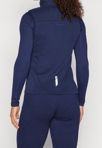 Námořnická sportovní bunda s dlouhými rukávy, vysokým límcem, hladkým povrchem a bílým páskem vzadu. Minimální viditelné branding.
