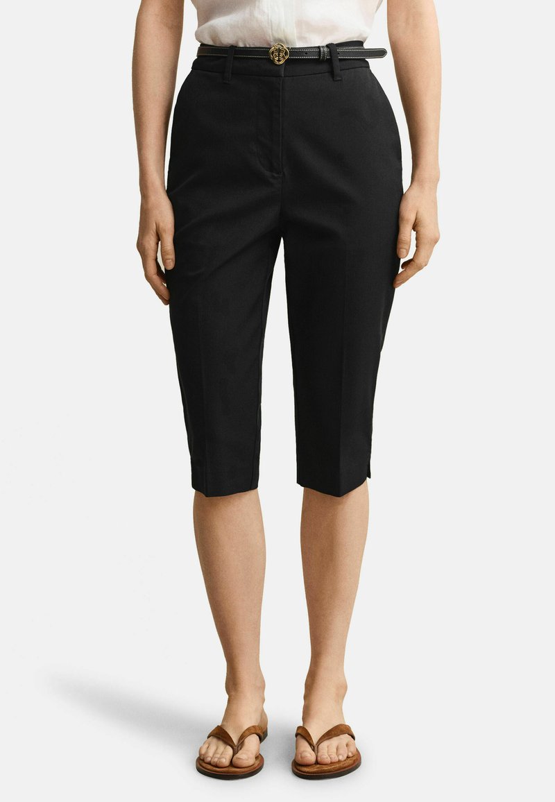 Femme portant un short noir ajusté longueur genou avec une ceinture noire et des sandales marron à brides, se tenant devant un fond blanc.