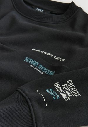 Svart sweatshirt med ribbad krage och ärmslut. Har vit och teal text, inklusive koordinater och fraser som "FRAMTIDENS SYSTEM" och "KREATIVA FRAMTIDSINDUSTRIER."