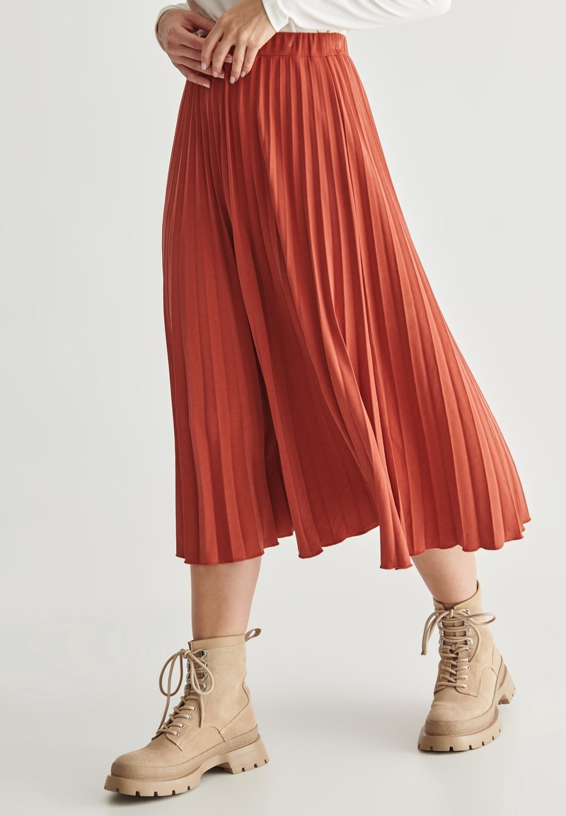 Faltenrock in Rostfarbe, aus weichem Material gefertigt. Kombiniert mit beigen Schnürstiefeln mit derben Sohlen.