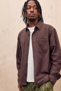 Camicia button-up a righe marroni con taschino e logo, indossata sopra una T-shirt bianca. Abbinata a pantaloni verdi con motivo.