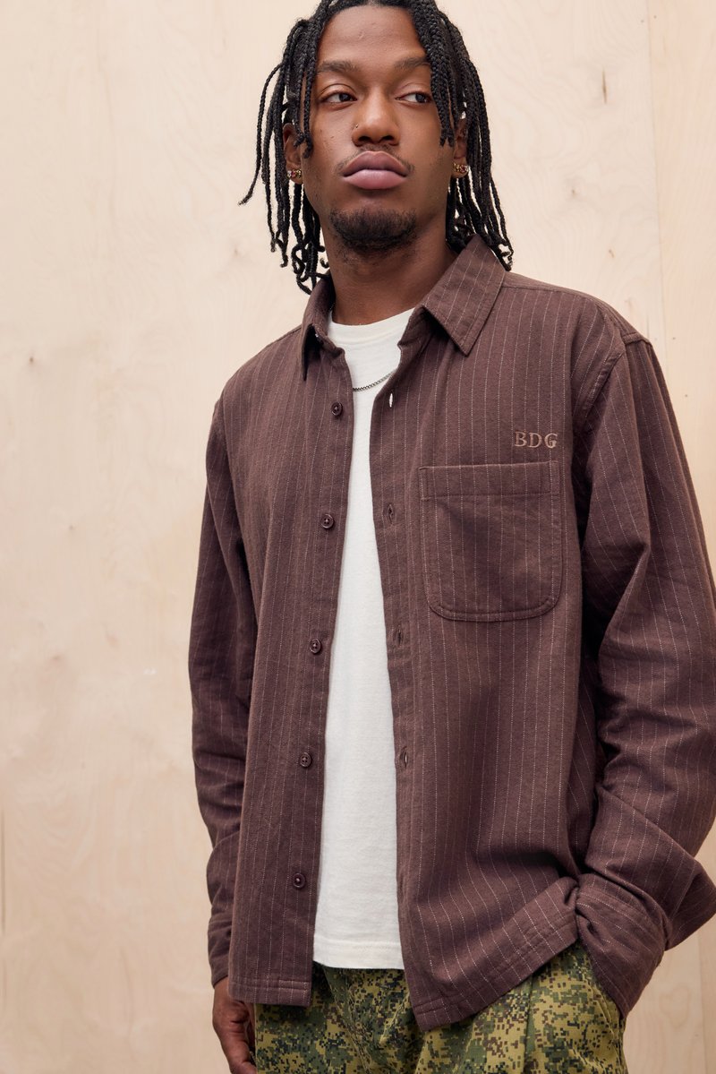 Camicia button-up a righe marroni con taschino e logo, indossata sopra una T-shirt bianca. Abbinata a pantaloni verdi con motivo.