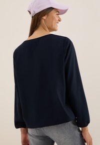 Camiseta de manga larga azul marino con un corte relajado, cuello redondo y mangas ligeramente fruncidas; combinada con jeans gris claro y una gorra rosa.