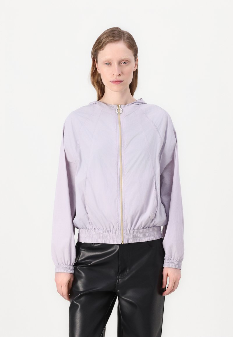 Licht lila jack met capuchon, volledige rits aan de voorkant, elastische manchetten en zoom. De stof lijkt glad en licht glanzend. Zwarte broek.