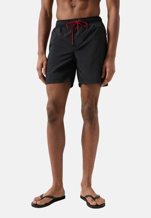 Mand iført sorte badeshorts med rød snøre og sorte klipklappere, stående mod en ensartet lys baggrund.