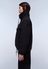 Mujer de perfil con una chaqueta negra de cuello alto y cordones ajustables, de pie frente a un fondo liso y claro.