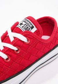 Zapatillas de lona rojas con una superficie texturizada, cordones blancos, ojales circulares y un parche de logo en blanco y negro en la lengüeta.