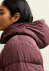 Chaqueta acolchada burdeos con cuello alto y capucha, presenta detalles de botones y una textura acolchada. Costuras visibles en el hombro.