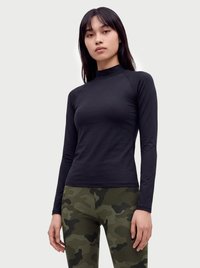 Jonge vrouw met een aansluitend zwart shirt met lange mouwen en groene camouflageleggings, staand met een neutrale uitdrukking tegen een effen achtergrond.