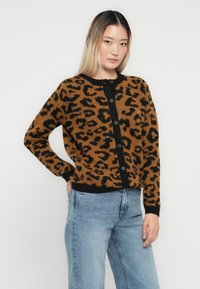 Cardigan en imprimé léopard brun et noir avec des boutons, col rond et poignets côtelés ; porté par-dessus un jean en denim bleu clair. Texture en tricot douce.