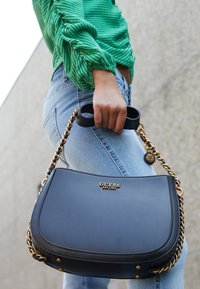 Osoba v modrých džínách a zelené blůze drží černou kabelku Guess se zlatým řetízkovým detailem na betonovém pozadí.